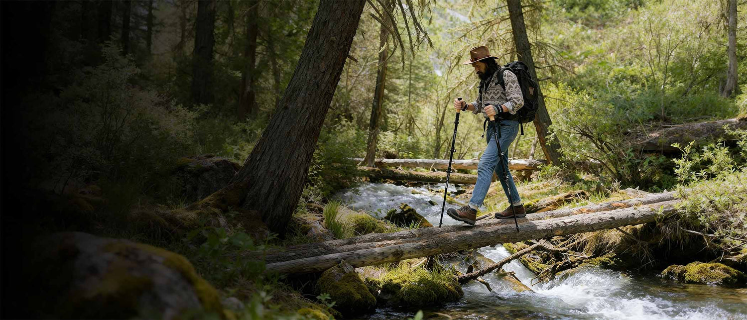 Person hiking through a forest with a backpack and walking sticks over a stream.