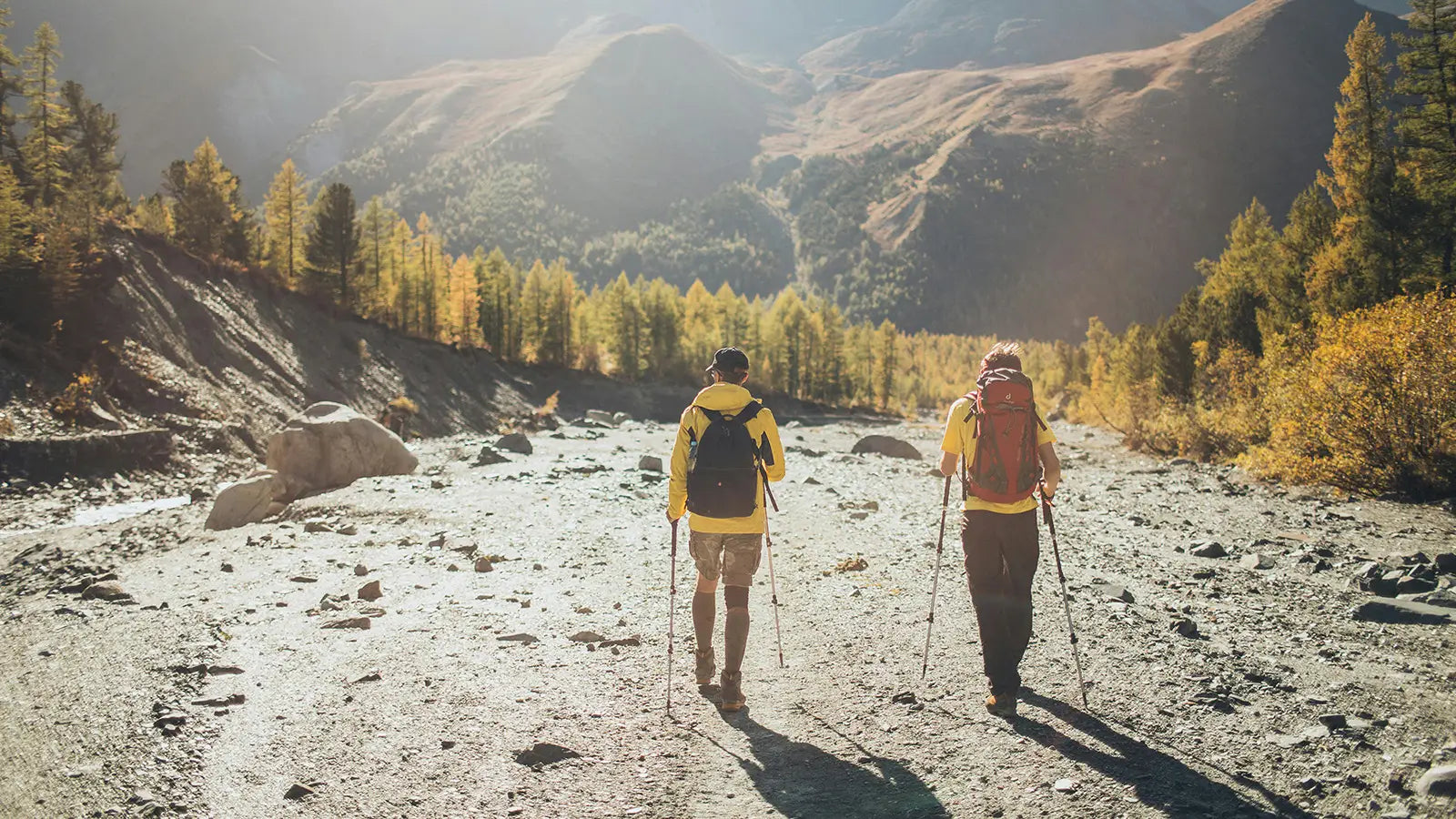 Two hikers with backpacks and trekking poles walking on a mountain trail with mountains and trees in the background.
